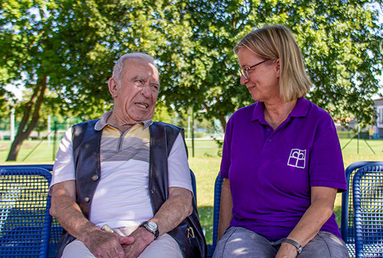 Betreuerin von der Tagespflege sitzt mit einem Senioren auf einer Parkbank.